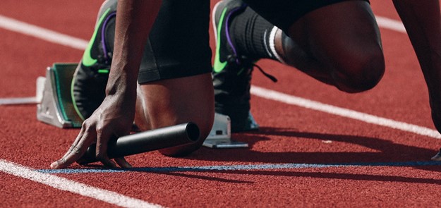De cerca la imagen de un atleta arrodillado en el punto de inicio de una pista de atletismo, con un bastón de relevo en la mano.