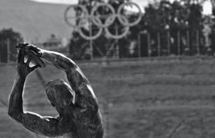 Imagen en blanco y negro de la espalda de una estatua de un atleta, con la escultura de los anillos olímpicos en la distancia.